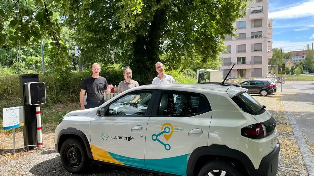 Ein Dacia Spring mit drei männlichenPersonen hinter dem Auto. Links ist eine Wallbox und im Hintergrund ein Baum sowie ein Gebäude.